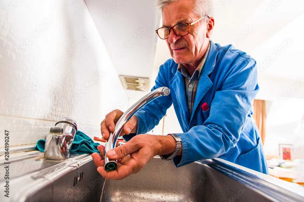 Older tradesman in blue overalls fixing a chrome kitchen tap over a sink