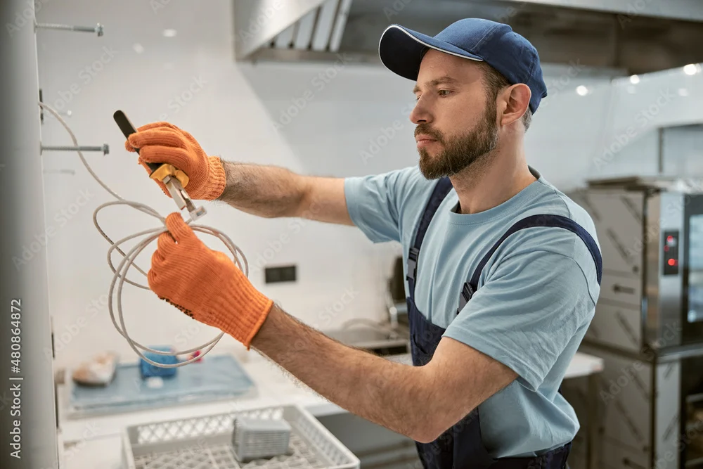 Electrician in a cap repairing an electric cable with pliers in a kitchen