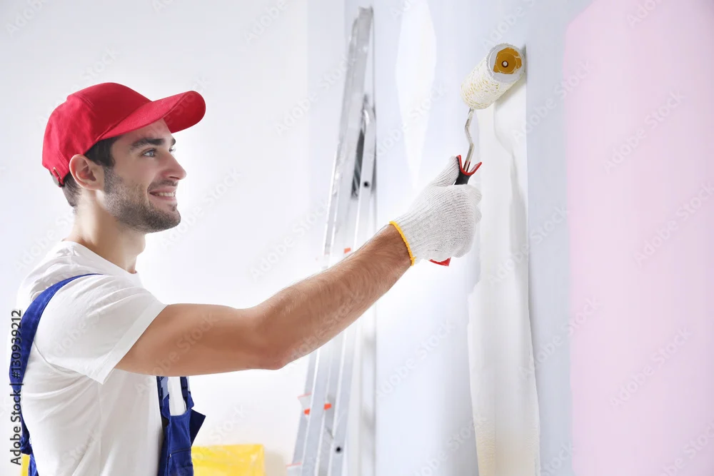 Smiling decorator in a red cap rolling paint on a wall beside a stepladder