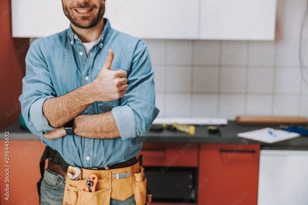 Smiling repairman wearing a tool belt and giving a thumbs up in a kitchen
